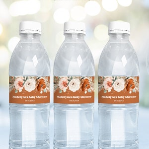 Może przedstawiać: Trzy przezroczyste plastikowe butelki z wodą z etykietami przedstawiającymi wzór kwiatowy w odcieniach brzoskwiniowego, białego i brązowego. Etykiety zawierają również tekst "Madelyne's Baby Shower" i datę "08.15.20XX".