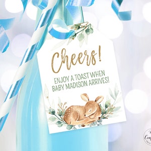 Puede incluir: Una etiqueta blanca con letras doradas que dice "Cheers! Enjoy a toast when baby Madison arrives!" La etiqueta está decorada con una ilustración de acuarela de un cervatillo durmiendo en una cama de hojas verdes.
