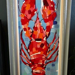 May include: A framed oil painting of a lobster. The lobster is painted in shades of red and orange against a blue background. The painting is in a rectangular, vertical format and is framed in a light gray frame.