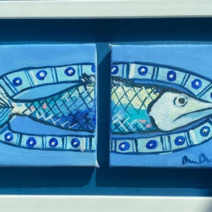 May include: A framed diptych painting of a fish. The fish is depicted in two parts, each on a square canvas. The fish is blue, white, and teal, with a decorative border. The artwork is signed.