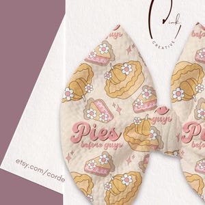 Könnte beinhalten: Eine Haarschleife aus Stoff mit einem wiederkehrenden Muster aus Kürbiskuchen und kleinen weißen Blumen auf cremefarbenem Grund. Der Text "Pies before guys" ist in einer rosa Retro-Schrift gedruckt. Die Schleife ist auf einer weißen Karte befestigt.