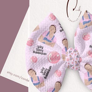 Puede incluir: Un lazo para el pelo de color rosa claro con una superficie de tela texturizada que presenta un patrón repetitivo de una persona con peto azul y camisa rosa soplando una gran burbuja rosa. El texto Icky Sticky Bubblegum está impreso en negro.