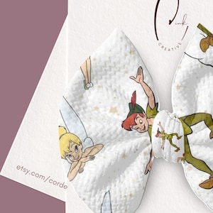 May include: A white hair bow featuring a repeating pattern of Peter Pan and Tinker Bell characters with small gold stars. The bow is attached to a white display card with the text Pink Creative and the URL etsy.com/cordercreativeink printed on it.