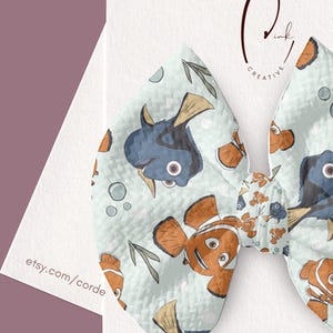 Peut inclure: Un nœud pour cheveux en tissu sur fond bleu clair avec un motif répétitif de poissons-clowns orange et de poissons chirurgiens bleus, agrémenté de petites bulles et d'algues vertes. Le nœud est fixé sur une carte blanche avec le texte etsy.com/cordercreativeink.
