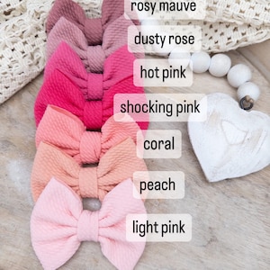 Peut inclure: Sept nœuds pour cheveux roses dans différentes nuances de rose, du rose clair au rose mauve. Les nœuds sont disposés verticalement, la nuance la plus claire en bas et la nuance la plus foncée en haut. Chaque nœud est étiqueté avec le nom de la nuance.