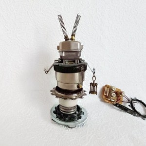 Op de afbeelding: Een zilveren en zwarte robot gemaakt van gerecyclede materialen. De robot heeft een rond hoofd met twee antennes, een rond lichaam met een tandwiel en een ronde basis. De robot staat op een wit oppervlak.