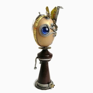 Puede incluir: Una escultura de estilo steampunk caprichosa hecha de un huevo, con un ojo de vidrio azul, acentos dorados y plateados y una cinta métrica amarilla. La escultura está montada sobre una base de madera marrón oscuro.