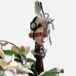 Puede incluir: Una escultura caprichosa hecha a partir de un huevo reutilizado, adornada con una correa de cuero, un reloj, gafas y plumas. El huevo está montado sobre una base de madera con un detalle de engranaje.