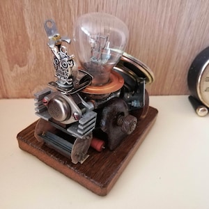 May include: A steampunk-style sculpture made from repurposed metal parts, including a light bulb, gears, and a small owl figurine. The sculpture is mounted on a wooden base.