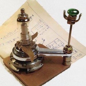 Könnte beinhalten: Eine einzigartige Steampunk-Skulptur mit einer Glasröhre, Metallkomponenten und einem bronzefarbenen Flügel. Die Skulptur ist auf einem braunen Holzsockel montiert, mit einem grünen Knopf und einem Metallarm. Im Hintergrund befindet sich ein Vintage-Papier mit Gleichungen.