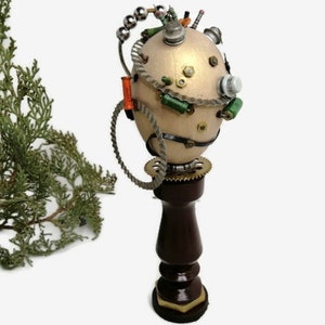Pode incluir: Escultura em estilo steampunk com uma forma ovóide bege claro adornada com vários componentes metálicos, incluindo engrenagens, fios e pequenos cilindros. Repousa sobre uma base de madeira torneada castanho escuro. Folhagem verde visível à esquerda.