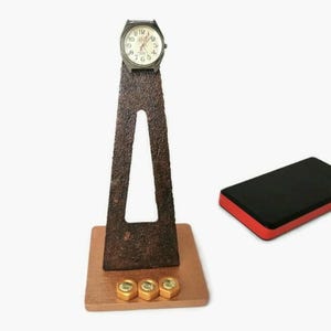 May include: A rustic brown metal watch stand with a triangular design. The stand has a small wooden base and four gold-colored nuts on the base. A silver watch is displayed on the stand. A red and black rectangular object is in the background.