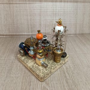 Puede incluir: Una escultura steampunk con engranajes, bombillas y otros componentes mecánicos. La escultura incluye un pequeño cilindro azul, esferas naranjas y un orbe blanco. Montado sobre una base cuadrada con un patrón de placa de circuito.