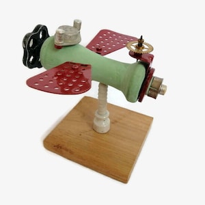 May include: A whimsical green and red aeroplane sculpture made from repurposed materials. The aeroplane has a propeller, wings, and a small wooden base.