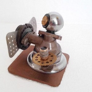May include: A silver and brass steampunk-style sculpture with a round, chrome ball on top, a gear, and a metal wing. The sculpture is mounted on a brown wooden base.