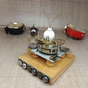 Op de afbeelding: Een steampunk sculptuur met een vacuümbuis, tandwielen en een kleine witte vaas met een koperdraad accent. De sculptuur is gemonteerd op een houten basis met metalen accenten. Andere vintage objecten zijn op de achtergrond.