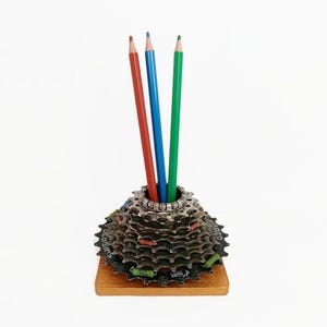 May include: A pencil holder constructed from a stacked metal bicycle cassette gear assembly mounted on a square wooden base. Three coloured pencils in red, blue, and green are inserted into the centre of the metallic, multi-layered gear structure.