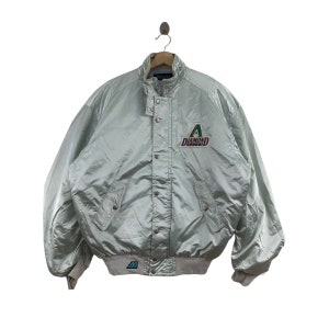 May include: A light silver satin jacket with a button-up front, two pockets, and a ribbed hem and cuffs. The jacket features a logo on the chest that says "Diamondbacks". The jacket is hanging from a hanger.