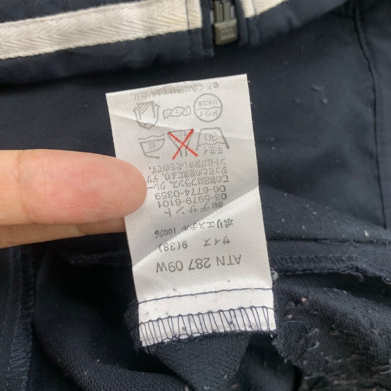 K&ouml;nnte beinhalten: Wei&szlig;es Etikett mit schwarzem Text und einem roten X &uuml;ber einem Waschsymbol. Der Text lautet "Dieses Produkt wurde in Frankreich von Descente in Zusammenarbeit mit Creuzet hergestellt."