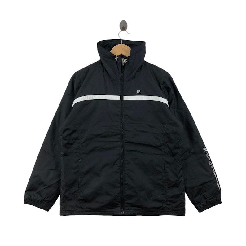 May include: Black athletic jacket with a high collar and a white horizontal stripe across the chest. The jacket has a full-length zipper and a small logo on the chest. The sleeves have text on the left sleeve. The jacket is hanging on a hanger.