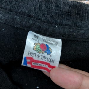 May include: A black Fruit of the Loom tag with white text that reads "Adult M 100% Cotton Preshrunk" and "Fruit of the Loom" with a red, white, and blue logo that reads "Made in U.S.A."