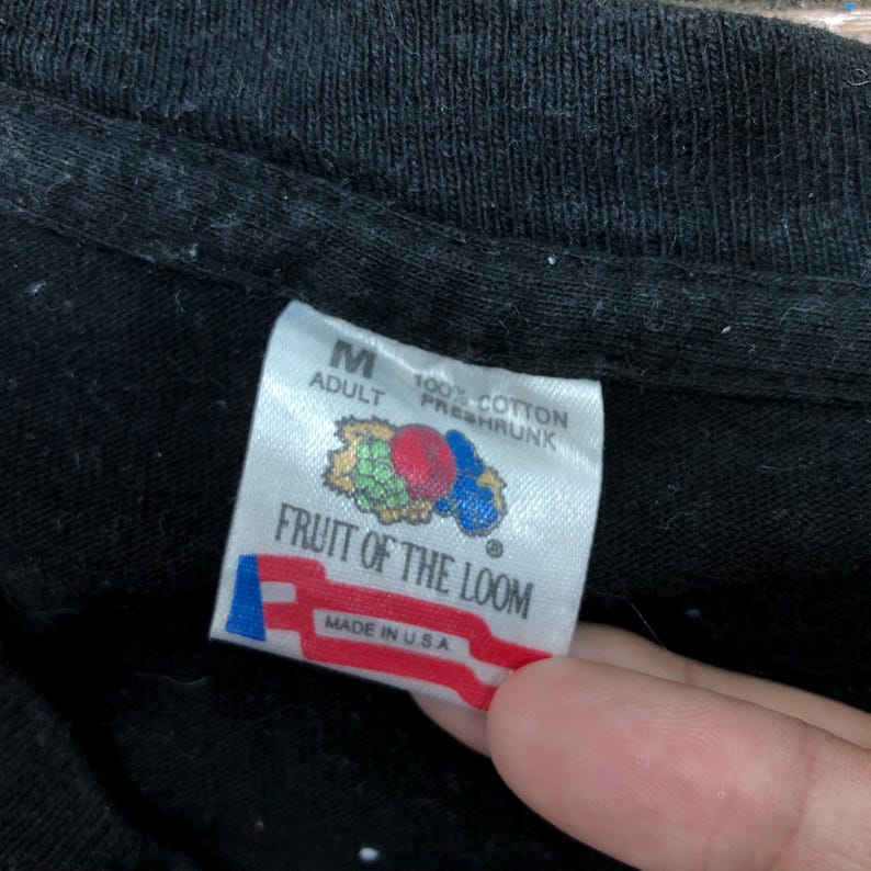 May include: A black Fruit of the Loom tag with white text that reads "Adult M 100% Cotton Preshrunk" and "Fruit of the Loom" with a red, white, and blue logo that reads "Made in U.S.A."