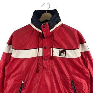 May include: Red and white Fila anorak jacket with a high collar and a small Fila logo. The jacket has a white stripe across the chest and a navy blue collar. Features a half-zip closure and two side pockets.