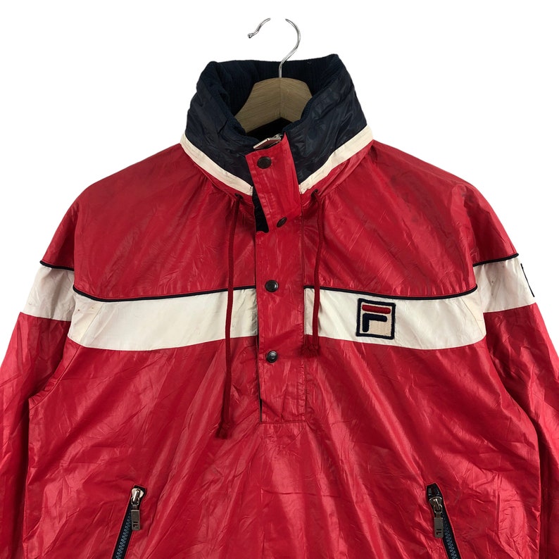 May include: Red and white Fila anorak jacket with a high collar and a small Fila logo. The jacket has a white stripe across the chest and a navy blue collar. Features a half-zip closure and two side pockets.