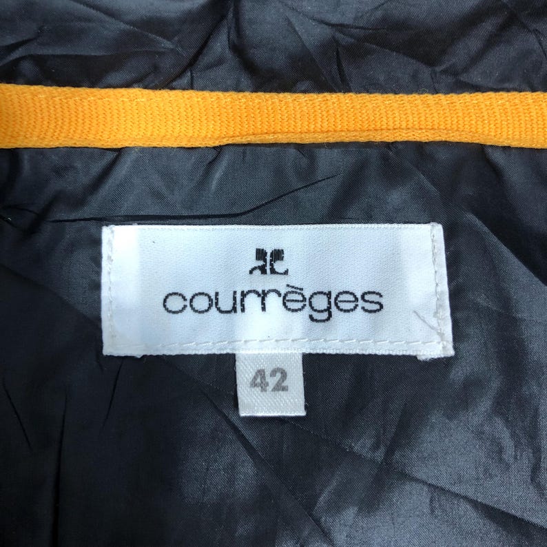May include: Close-up of a black garment with a yellow trim. A white label with the brand name "courr&egrave;ges" and the number "42" is visible. The garment appears to be made of a smooth, possibly synthetic material.