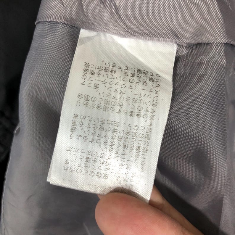 May include: Close-up of a white clothing tag with Japanese text, attached to a gray garment. The tag appears to provide care instructions and other details about the item. The fabric of the garment is visible in the background.