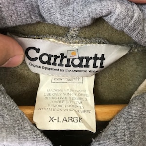 以下が含まれることがあります： グレーの Carhartt パーカー。白いタグには「Carhartt Original Equipment for the American Worker」と記載されており、小さなタグには洗濯方法とサイズ「X-Large」が記載されています。