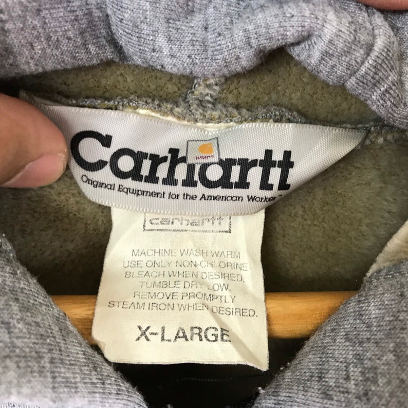 以下が含まれることがあります： グレーの Carhartt パーカー。白いタグには「Carhartt Original Equipment for the American Worker」と記載されており、小さなタグには洗濯方法とサイズ「X-Large」が記載されています。