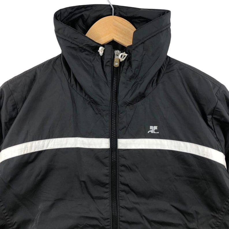 May include: Black jacket with a white stripe across the chest. The jacket has a high collar and a full-length zipper. The jacket is hanging on a wooden hanger. A small logo is visible on the right chest.