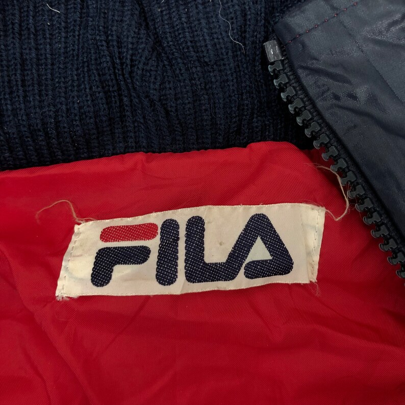May include: Close-up of a navy blue jacket with a red lining. The jacket features a white label with the brand name "FILA" in navy blue letters and a red stripe. A black zipper is visible.