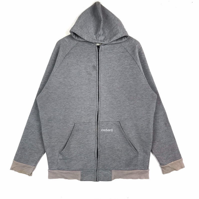 以下が含まれることがあります： 前面に Carhartt のロゴが付いたグレーのジップアップフーディー。フードにはドローストリングが付いており、カンガルーポケットが付いています。