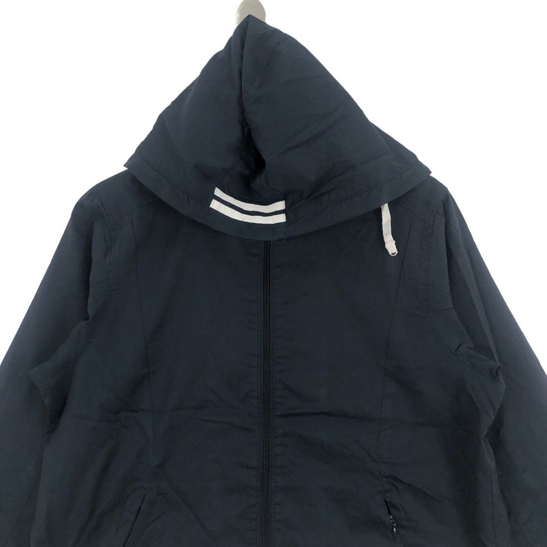 K&ouml;nnte beinhalten: Eine marineblaue Kapuzenjacke mit einem wei&szlig;en Streifen-Detail an der Kapuze. Die Jacke hat einen Rei&szlig;verschluss und eine Kapuze mit Kordelzug.