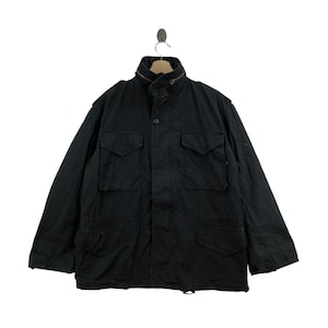 May include: Black military-style jacket with four front pockets and a high collar. The jacket has a button-up front and a drawstring at the bottom. The jacket is hanging on a metal hook.