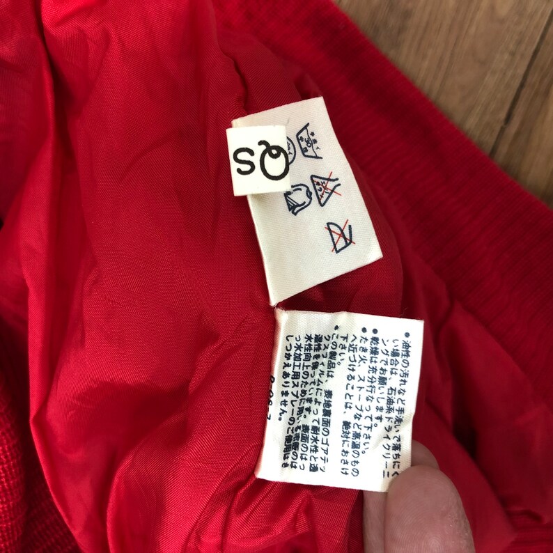May include: Close-up of a red garment's interior, revealing a red lining and a white tag with the letters "SQ" and care symbols. Another tag with Japanese text is also visible.