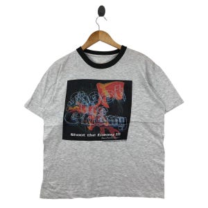 May include: A gray ringer t-shirt with a black collar and cuffs. The shirt features a graphic with the text "Shoot the Enemy!!!" in a retro style, with red and blue accents.