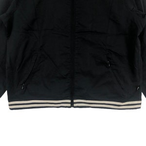 K&ouml;nnte beinhalten: Schwarze Rei&szlig;verschlussjacke mit einem wei&szlig;en und schwarzen Streifenbesatz am unteren Rand.