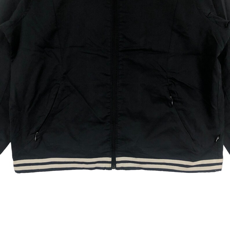 K&ouml;nnte beinhalten: Schwarze Rei&szlig;verschlussjacke mit einem wei&szlig;en und schwarzen Streifenbesatz am unteren Rand.