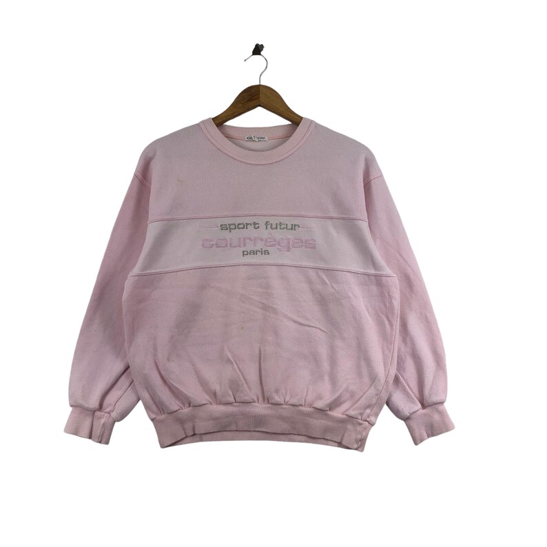 May include: A light pink Courreges Paris sweatshirt. The sweatshirt has a crew neck and long sleeves. The words "sport futur" and "courreges paris" are embroidered in a lighter pink across the chest. The sweatshirt is hanging on a wooden hanger.