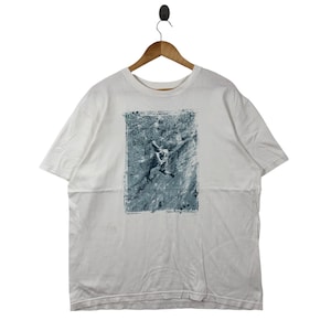 May include: White t-shirt with a graphic of a person climbing a rock face. The image is in shades of gray and blue, framed by a rectangular border. The shirt is hanging on a wooden hanger.
