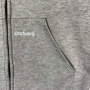 以下が含まれることがあります： 左胸ポケットに刺繍されたCarharttロゴが付いたグレーのジッパー付きフーディーのクローズアップ。
