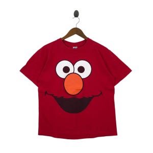 May include: Red t-shirt featuring a large graphic of a cartoon character's face. The face has large white eyes, an orange nose, and a smiling mouth. The shirt is hanging on a wooden hanger.