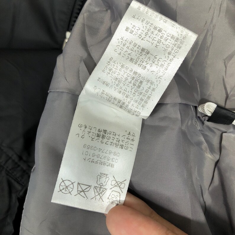May include: Close-up of a clothing label with Japanese text and care symbols. The label is white with black text and is attached to a gray lining. The label provides washing instructions and company information.