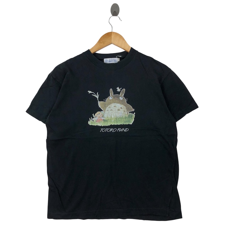 May include: Black t-shirt featuring a graphic of Totoro from the animated film My Neighbor Totoro. The design includes Totoro, a small girl, and the text "TOTORO FUND" in white. The shirt is hanging on a wooden hanger.