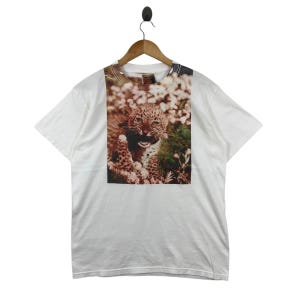 Può includere: T-shirt bianco con una grande grafica di un cucciolo di leopardo che ringhia in mezzo a fiori e verde. Il leopardo ha macchie marroni ed è ambientato su uno sfondo color seppia. La maglietta è appesa a una gruccia di legno.
