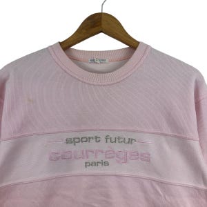 May include: A light pink sweatshirt with the text "sport futur courr&egrave;ges paris" embroidered in silver thread. The sweatshirt is hanging on a wooden hanger.