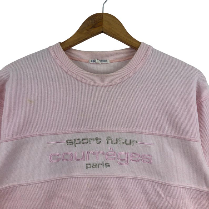 May include: A light pink sweatshirt with the text "sport futur courr&egrave;ges paris" embroidered in silver thread. The sweatshirt is hanging on a wooden hanger.